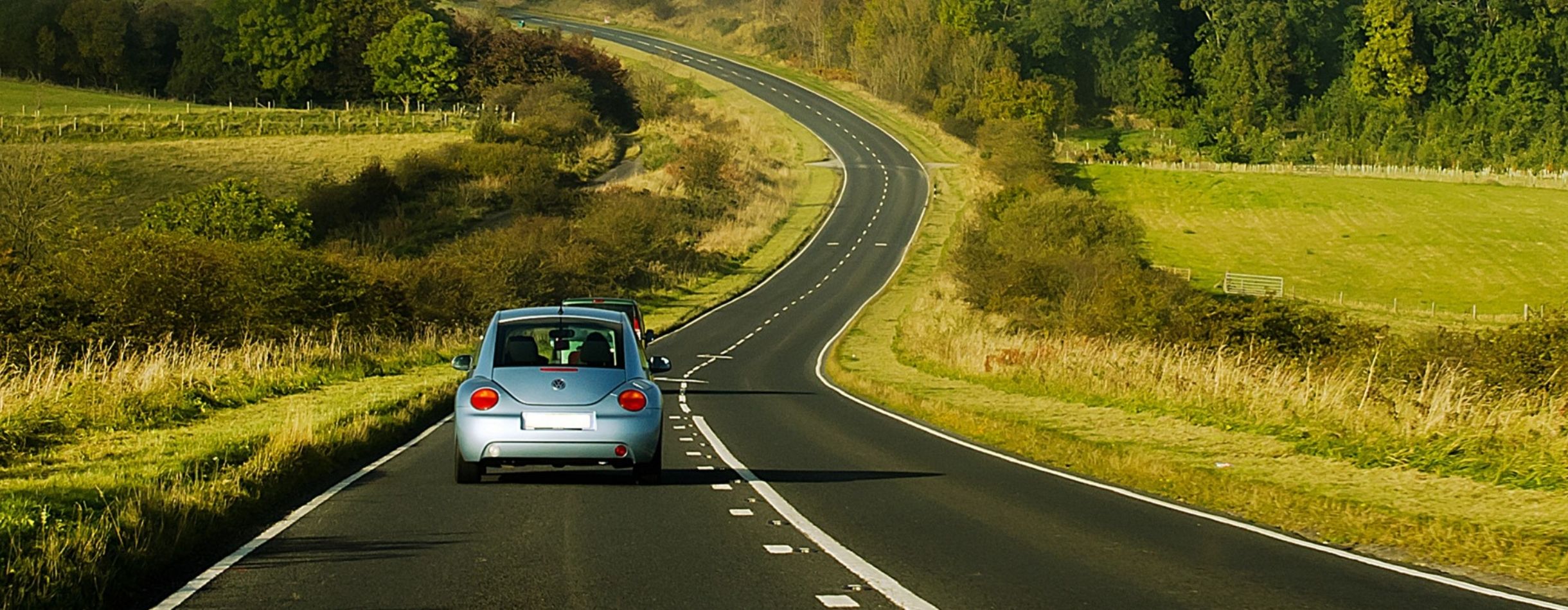 Two small cars traveling along an empty road with grass fields and short tree lines on each side.