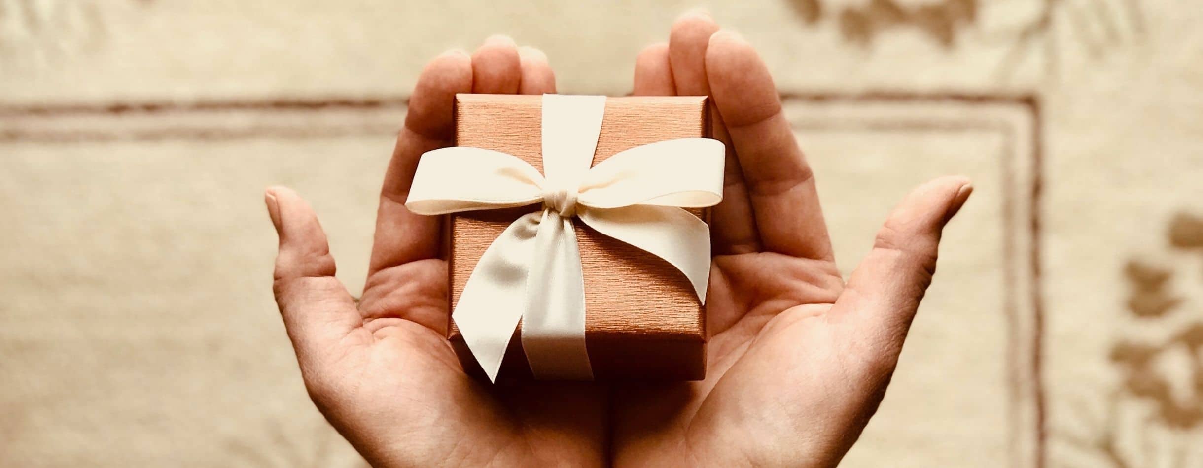 A small orangish-red gift that's wrapped in a tan colored bow lays delicately between two extended hands of a person.