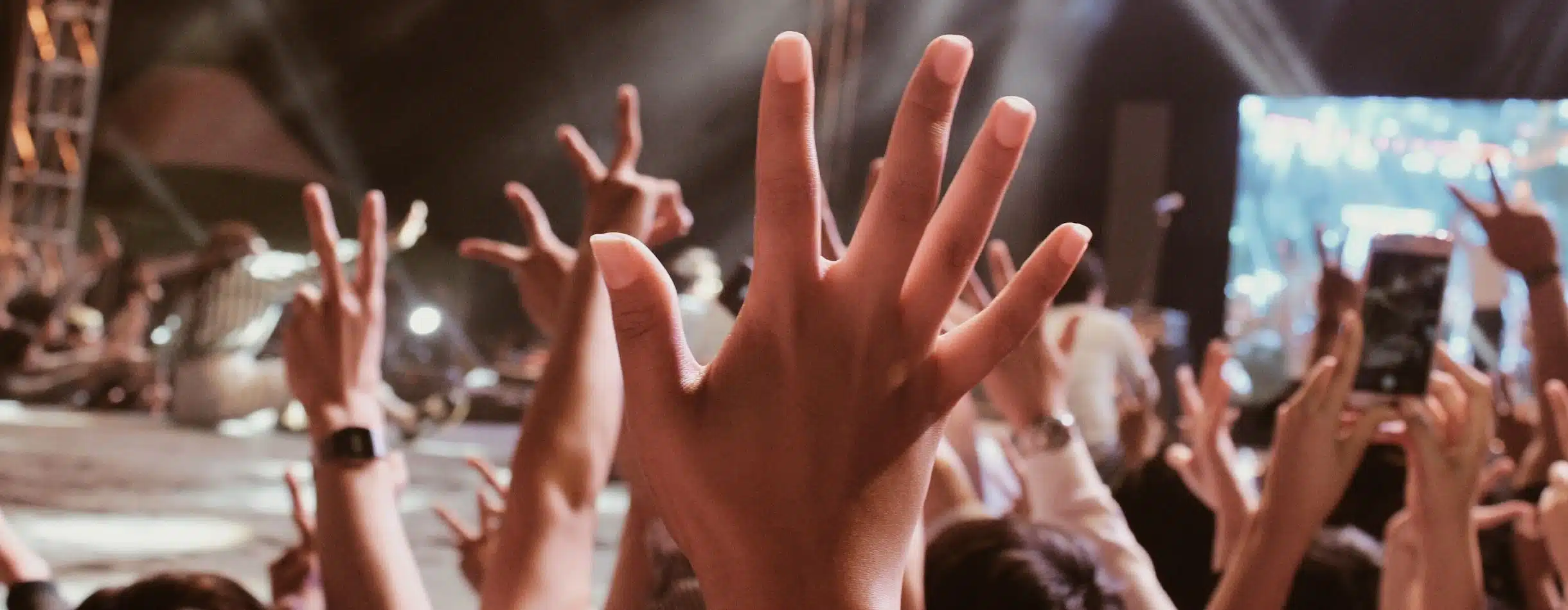 Several people have their hands raised at a concert, one person is filming with their phone, there is an infectious energy in the crowd