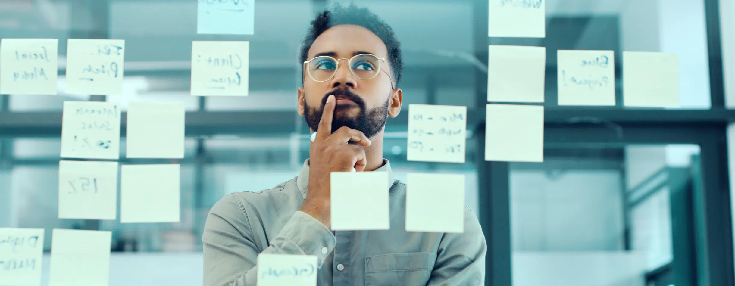 A man ponders at a wall of sticky notes. He rests his index finger on his upper lip as he brainstorms.
