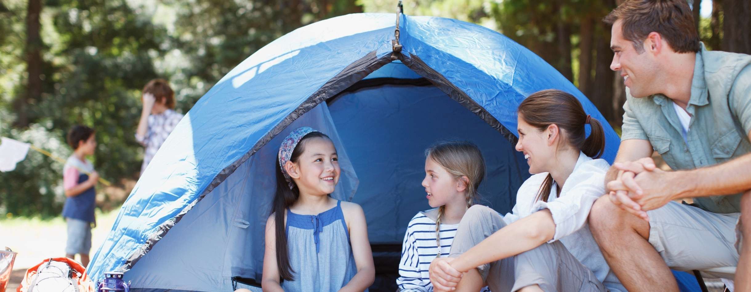 A young family is having a conversation outside their medium-sized blue tent. In the background, there's a kid with a bug net.