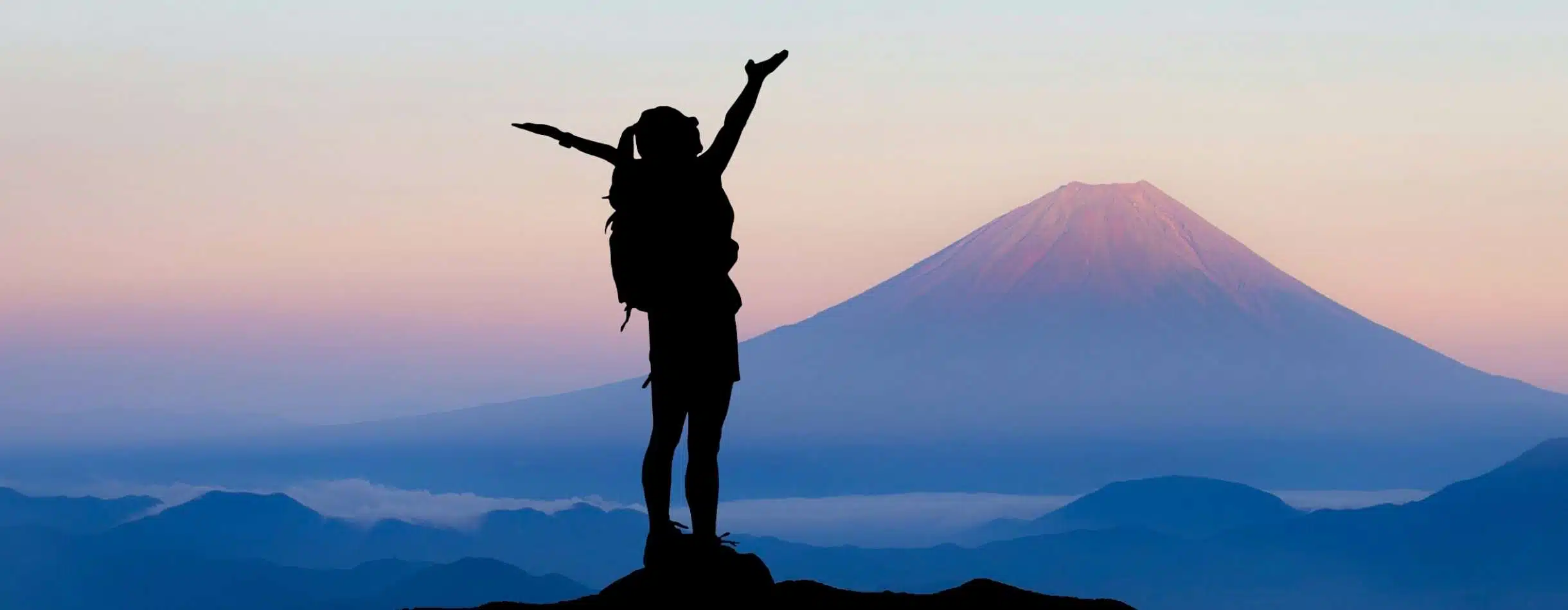 A woman extends her arms towards the sky in relief as they summit a smaller mountain with mount Kilimanjaro in the distance.
