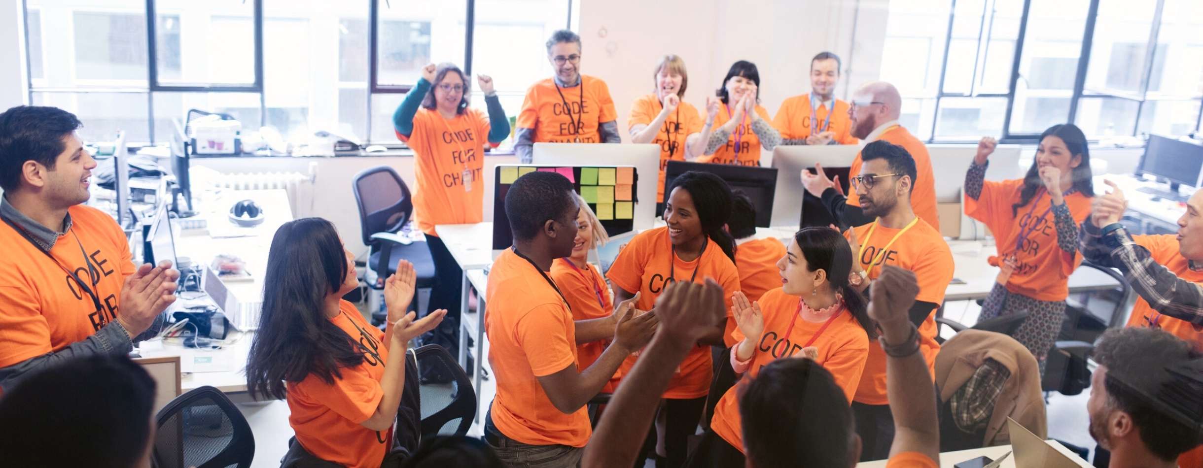 A large group of volunteers and nonprofit professionals celebrate after a successful fundraising round in their office. Everyone has matching orange shirts on.
