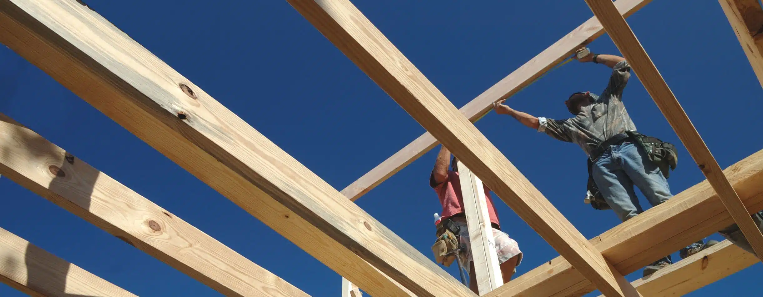 Two construction workers stand on wooden beams as they continue to assemble a house's frame.