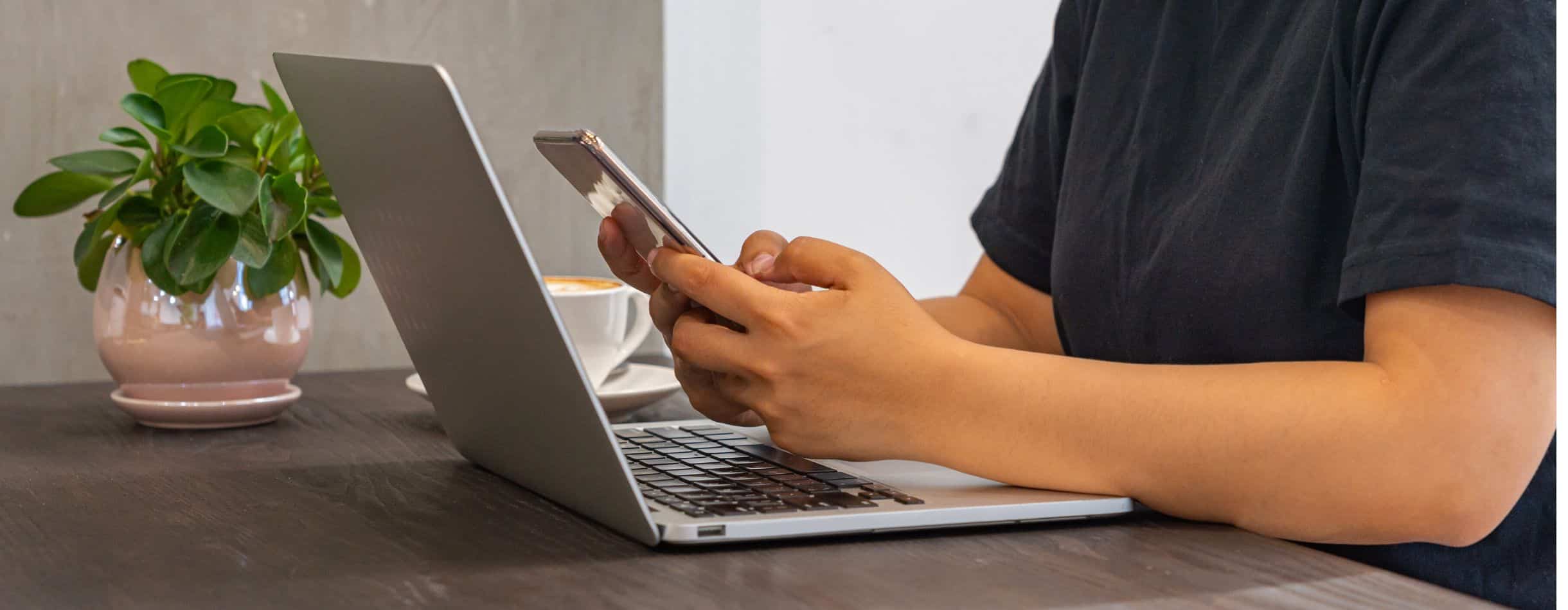 A person is working on their laptop but instead of typing on the keyboard, they're using their phone to text - likely reaching out to a donor for their nonprofit.