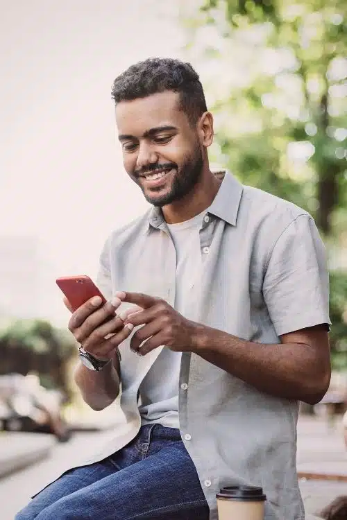A man smiles as he looks down at his iphone. He's wearing an open collar short sleeve button up shirt with blue jeans and is sitting outside in a park.