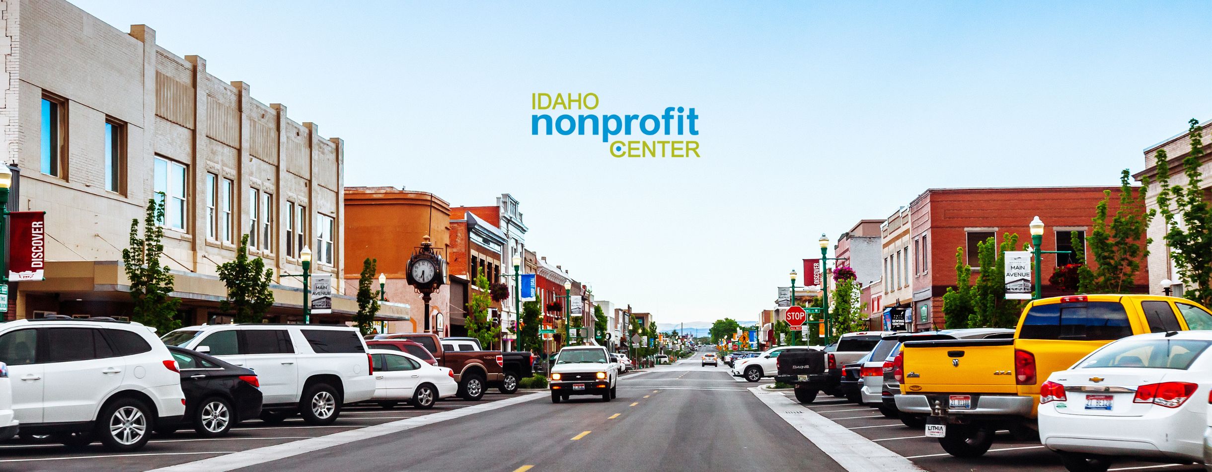 A downtown area in Idaho is shown with various cars parked on the and people shopping at nearby businesses. The Idaho Nonprofit Center Logo is displayed at the top of the image, with the sky being its backdrop.