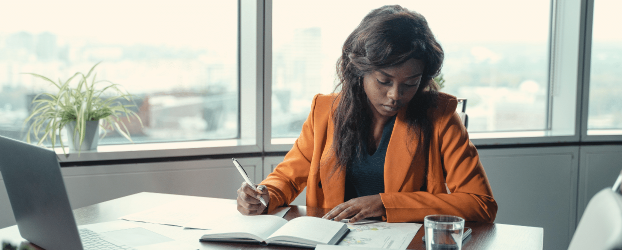 A woman in an orange jacket works on a development plan at her office desk.